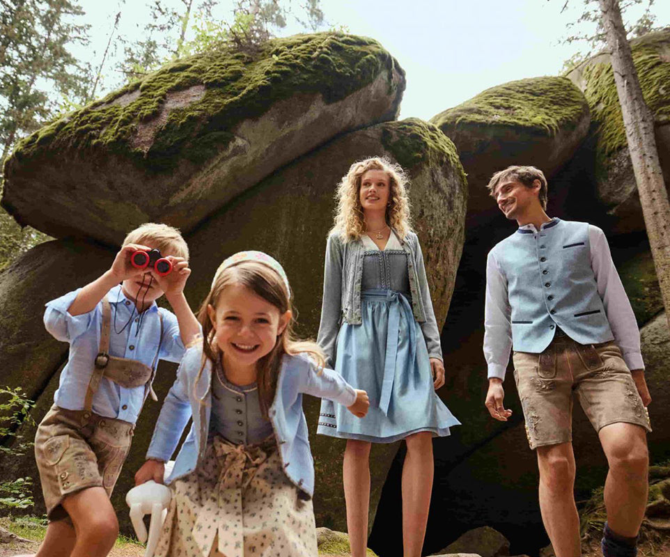 2 children and 2 parents in traditional Bavarian clothing standing in a forest with large rocks and trees.